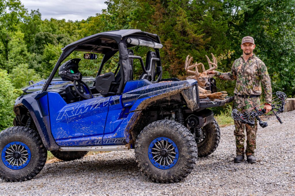Hunter standing next to Side-by-Side with deer in the back